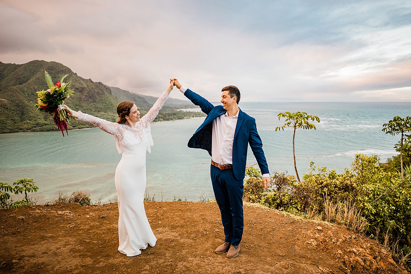 Adventurous Oahu elopement. The bride and groom holding hands celebrating on a cliff in front of the ocean.
